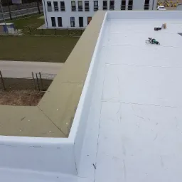 Płaski dach pokryty białą membraną, widoczne narzędzia do zgrzewania i docieplenie krawędzi dachu z widocznymi mocowaniami, budynek w tle.