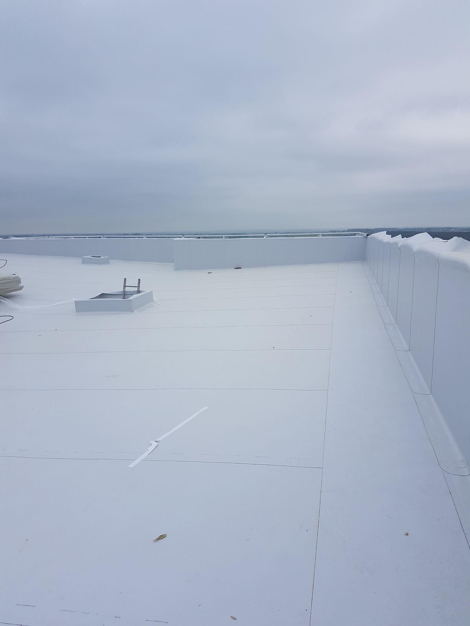 Jasny, płaski dach pokryty białą membraną dachową z widocznymi detalami, takimi jak kominki wentylacyjne i zabezpieczenia krawędzi, na tle pochmurnego nieba.