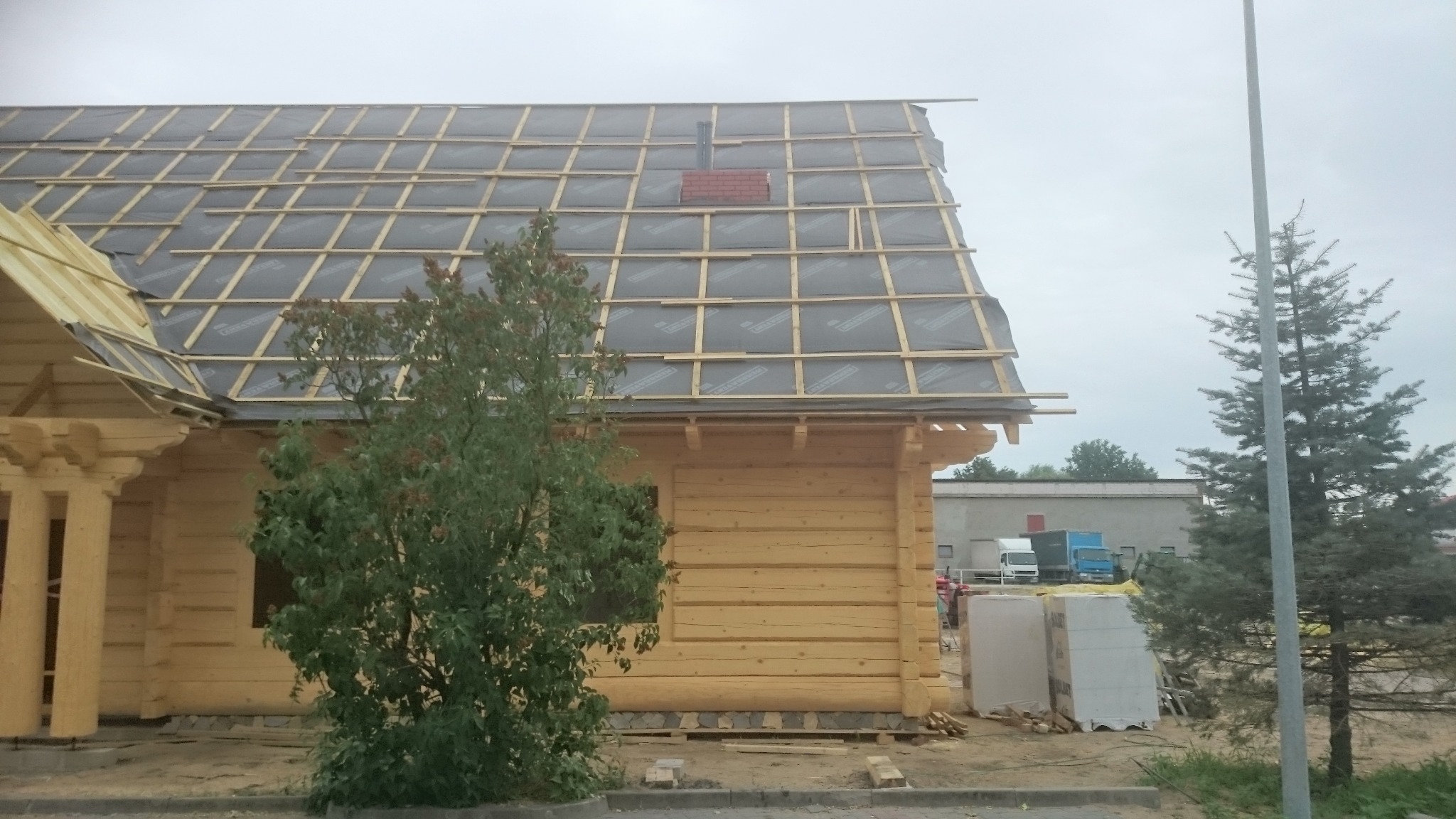 Drewniany dom w trakcie budowy, widoczna konstrukcja dachu z membraną, łatami i fragmentem komina z cegły. Na pierwszym planie drzewo liściaste, w tle budynek gospodarczy i samochody dostawcze.