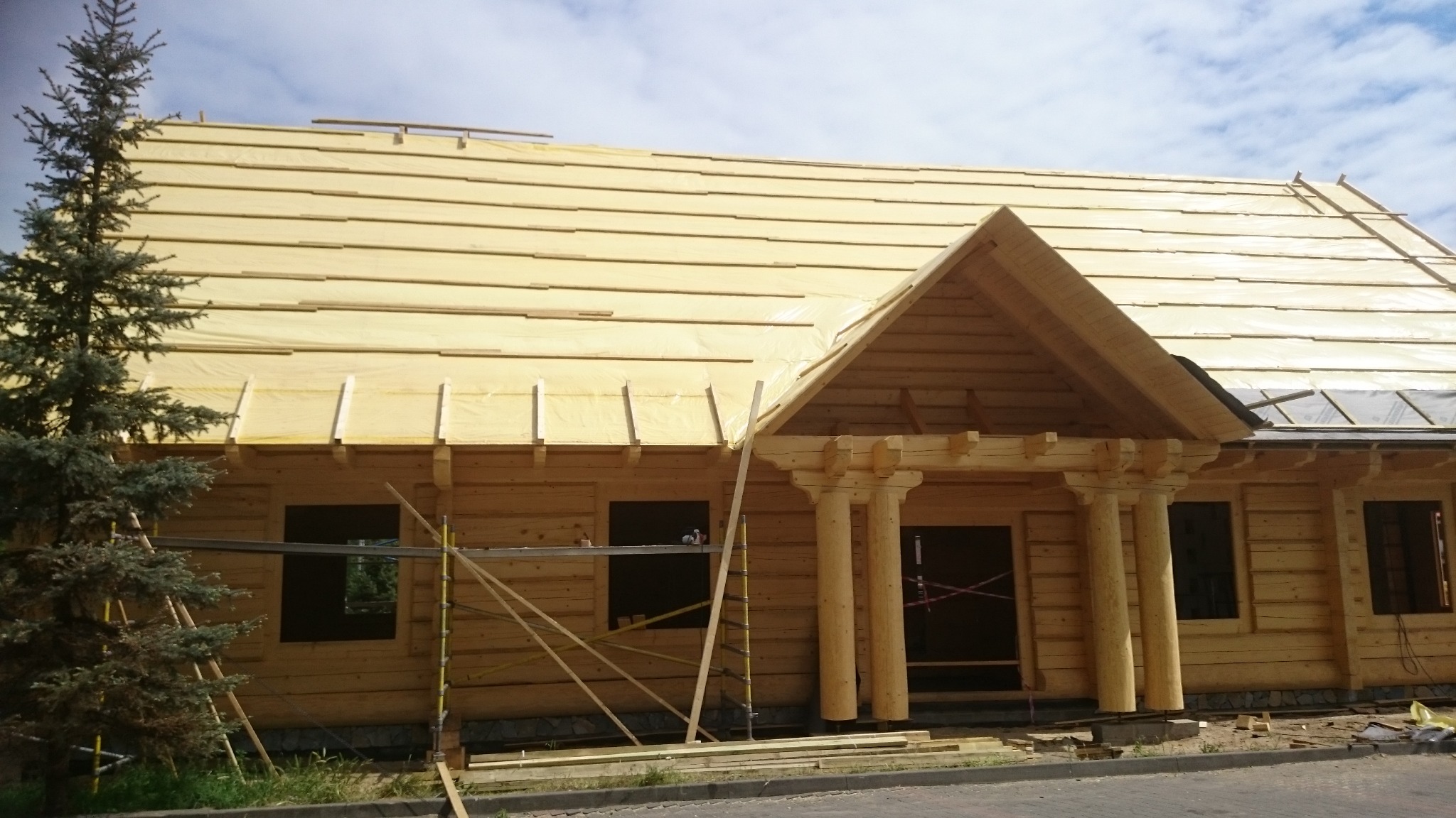 Surowy drewniany dom w trakcie budowy, z widocznym dachem pokrytym żółtą folią ochronną i elementami konstrukcyjnymi, rusztowania przy ścianach, świerk przed budynkiem.
