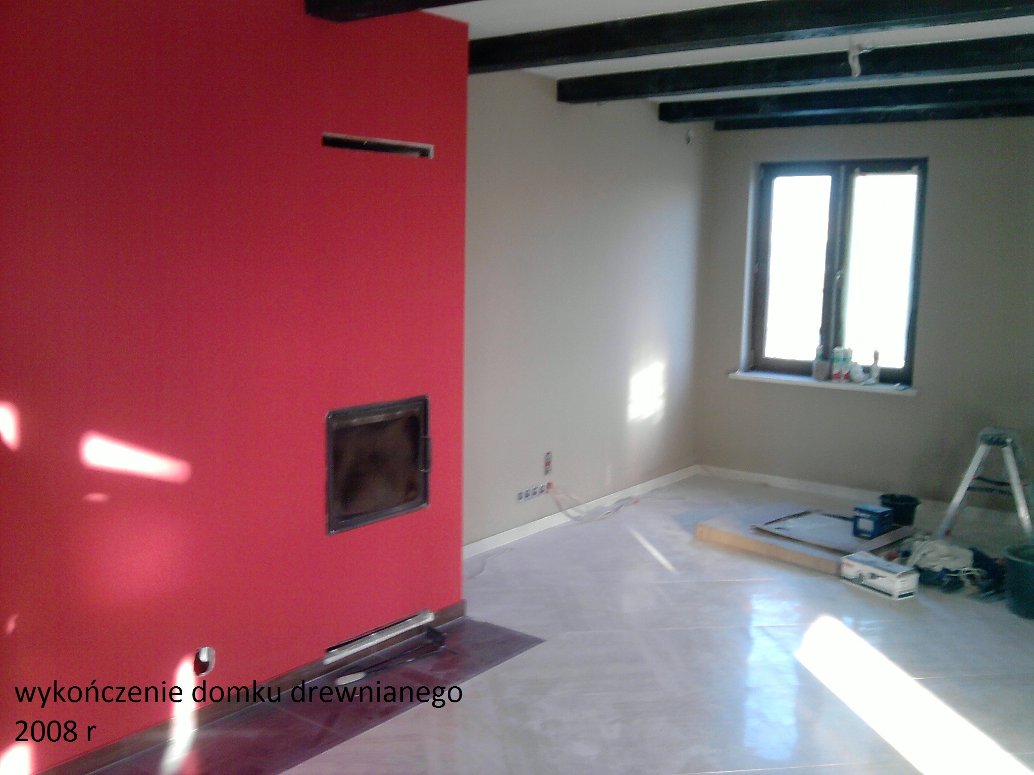 Wnętrze w trakcie wykańczania drewnianego domu: czerwona ściana z wkładem kominkowym, surowe drewniane belki sufitowe, okno z butelkami na parapecie, zabezpieczona podłoga i drabina w rogu.