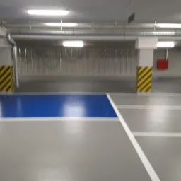 Posadzka żywiczna w garażu podziemnym z wyznaczonym niebieskim miejscem parkingowym i żółto-czarnymi pasami na słupach.