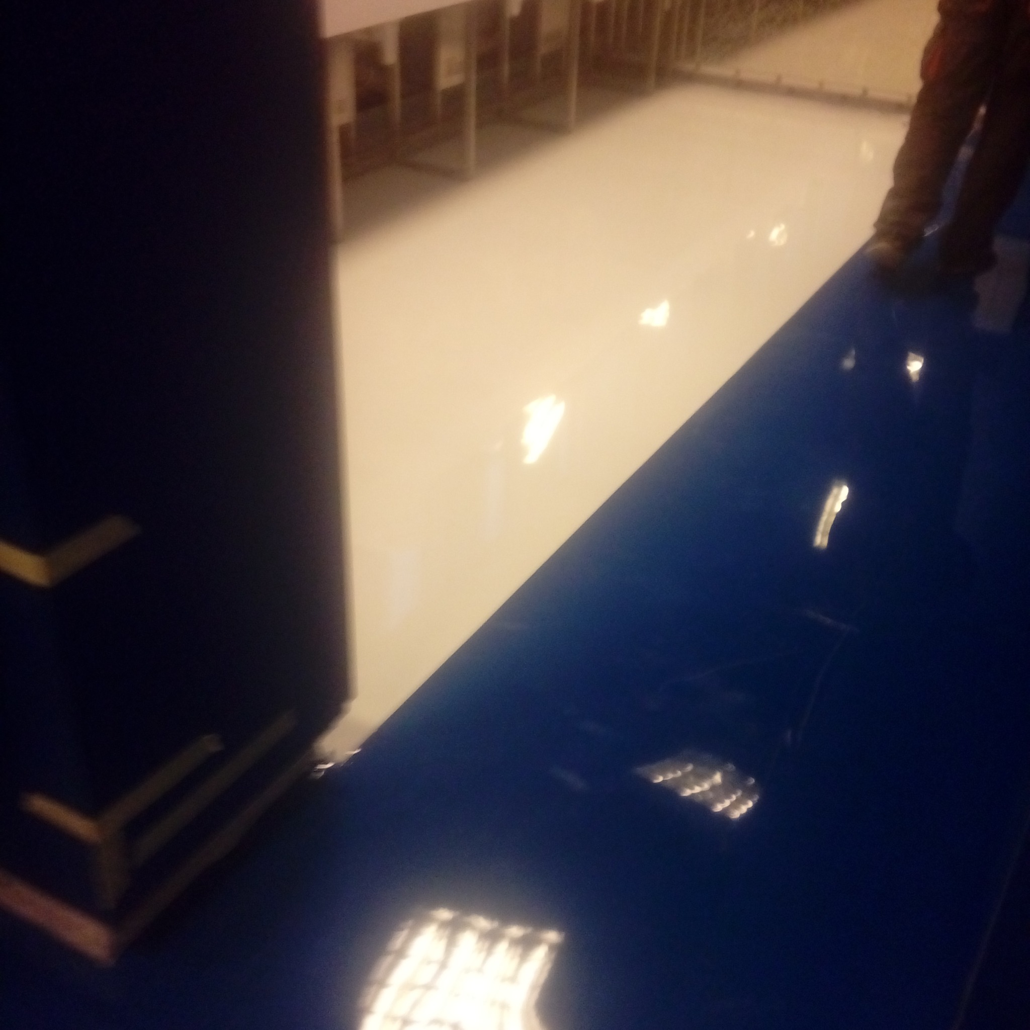 Biała i niebieska, błyszcząca podłoga w hali, widoczne nogi osoby w roboczych butach, fragment regału w tle.