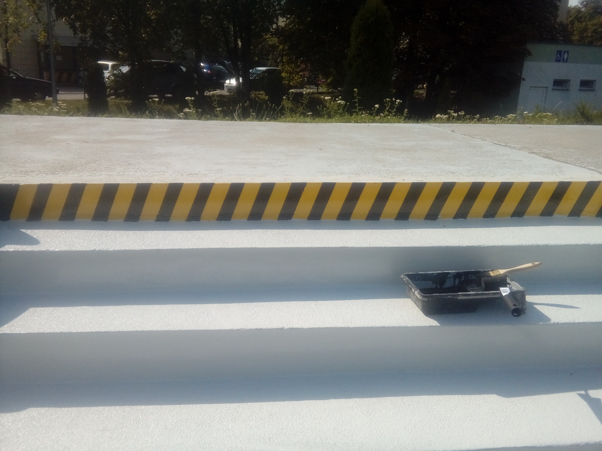 Świeżo pomalowane, białe schody zewnętrzne z żółto-czarnym pasem ostrzegawczym na górnej krawędzi, wiaderko z farbą i pędzel na jednym ze stopni.