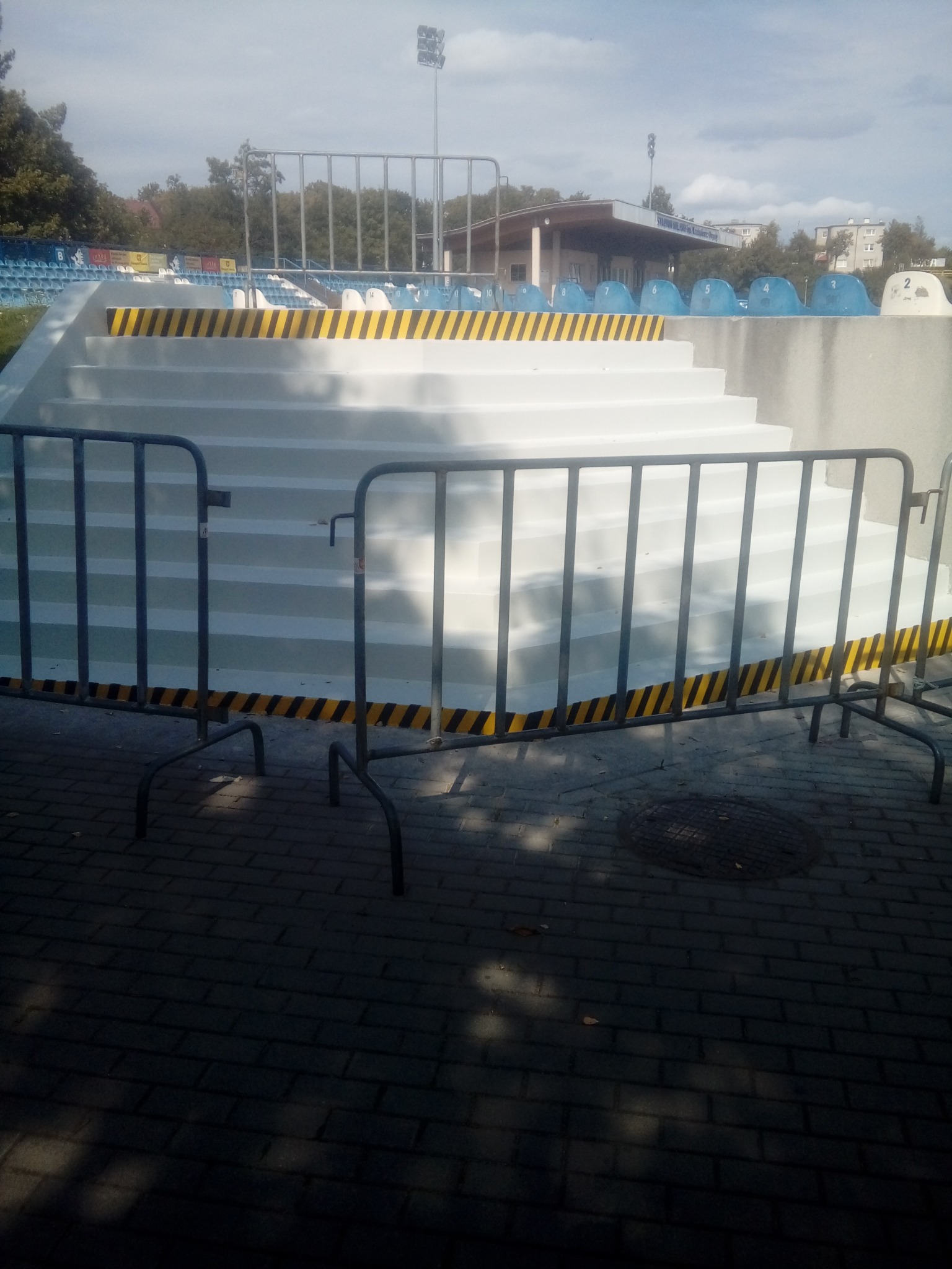 Białe, odnowione schody na stadionie z żółto-czarnymi pasami ostrzegawczymi, zabezpieczone metalowymi barierkami ochronnymi.
