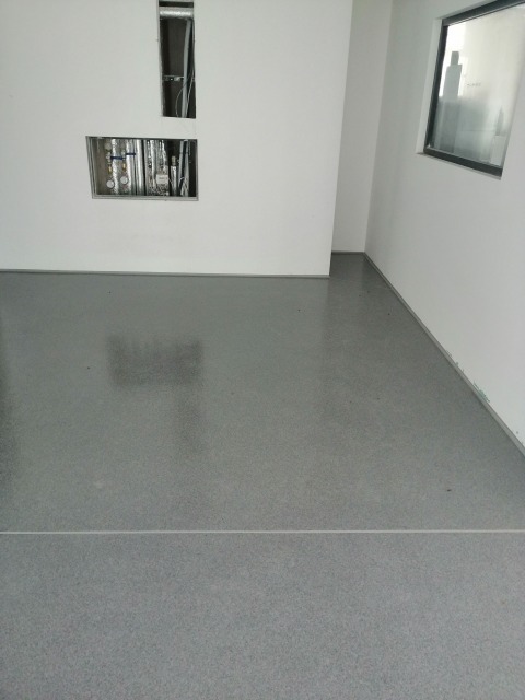 Szara, gładka posadzka w jasnym pomieszczeniu z wnękami na instalacje i oknem. Widoczna linia podziału na posadzce.