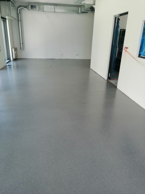 Szara, gładka posadzka żywiczna w jasnym, pustym pomieszczeniu z widocznymi elementami wentylacji pod sufitem i przygotowanym otworem drzwiowym.
