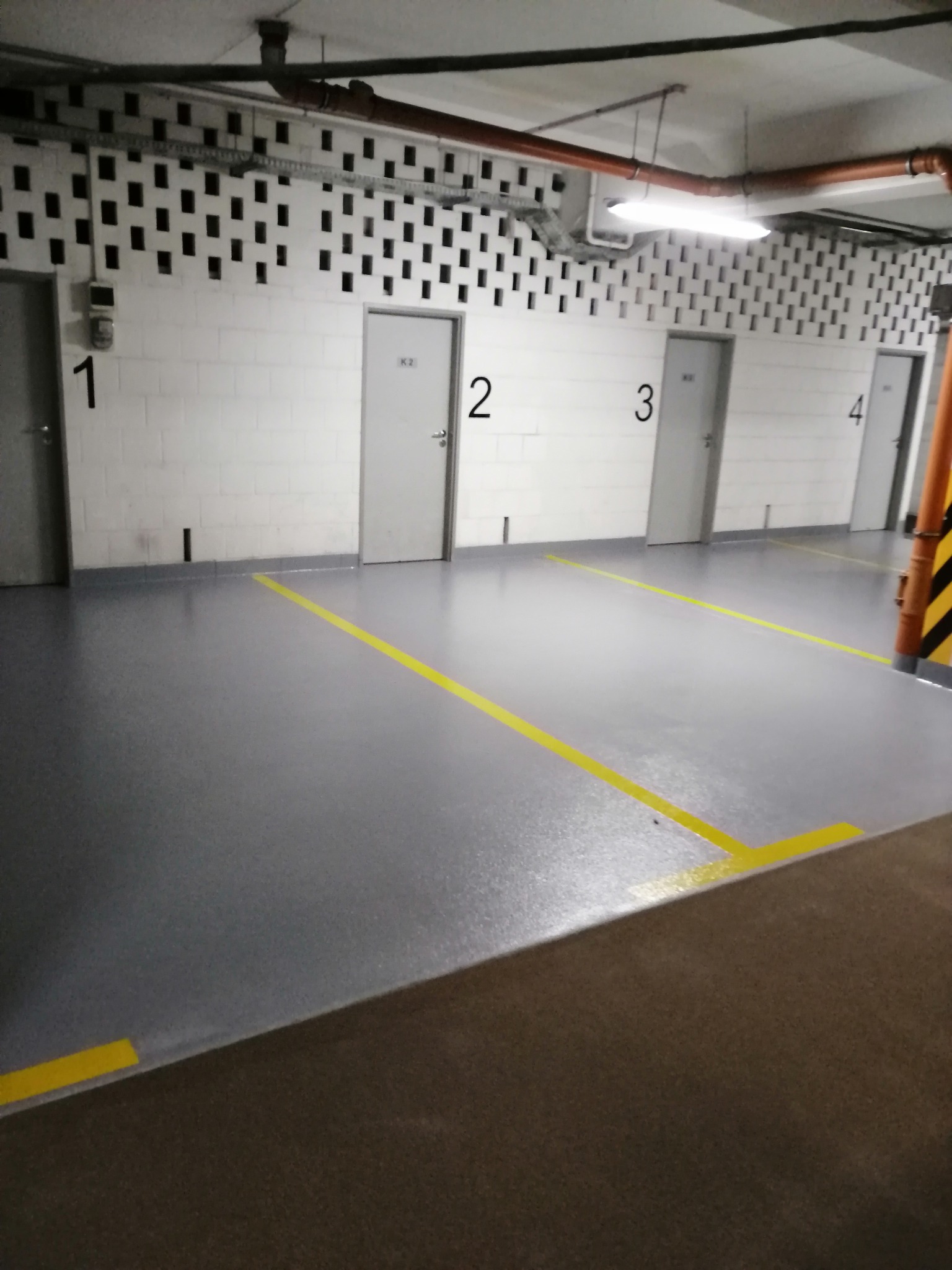 Szara podłoga w garażu podziemnym z żółtymi liniami wyznaczającymi miejsca parkingowe, widoczne cztery białe drzwi z numerami 1-4, ściany z białej cegły z otworami wentylacyjnymi pod sufitem.