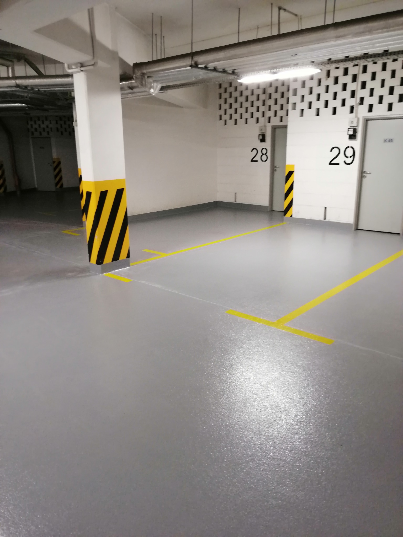 Szara posadzka w garażu podziemnym z żółtymi oznaczeniami miejsc parkingowych i słupami w pasy ostrzegawcze.