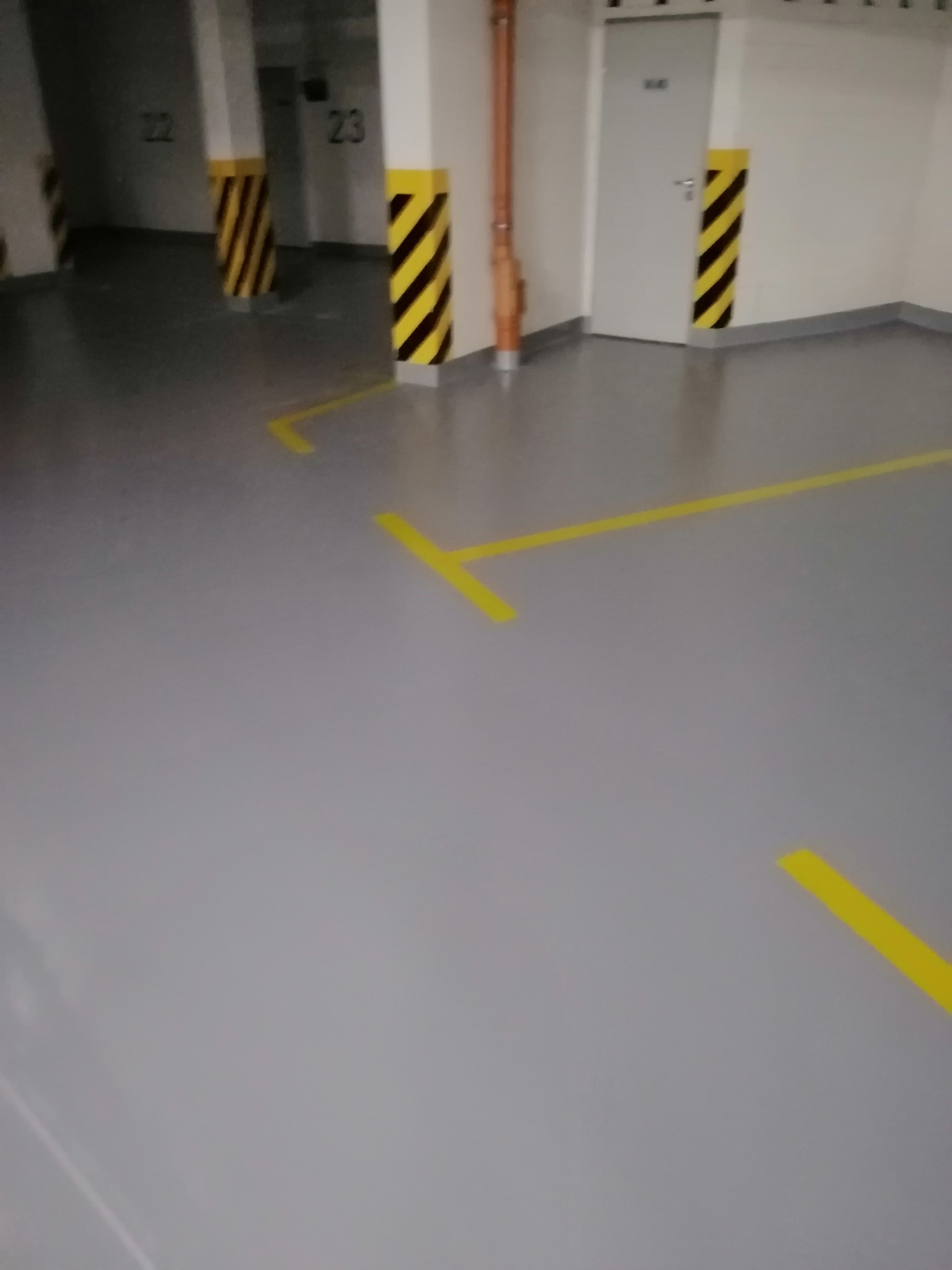 Szara posadzka w garażu podziemnym z żółtymi liniami wyznaczającymi miejsca parkingowe, słupy oznaczone czarno-żółtymi pasami ostrzegawczymi.