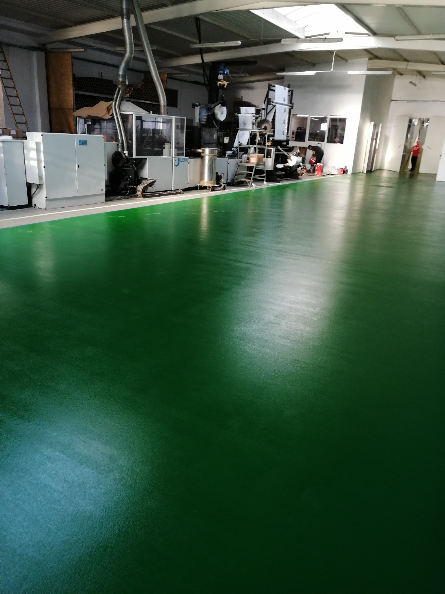 Błyszcząca, jednolita, zielona posadzka w hali produkcyjnej z maszynami i pracownikami w tle.