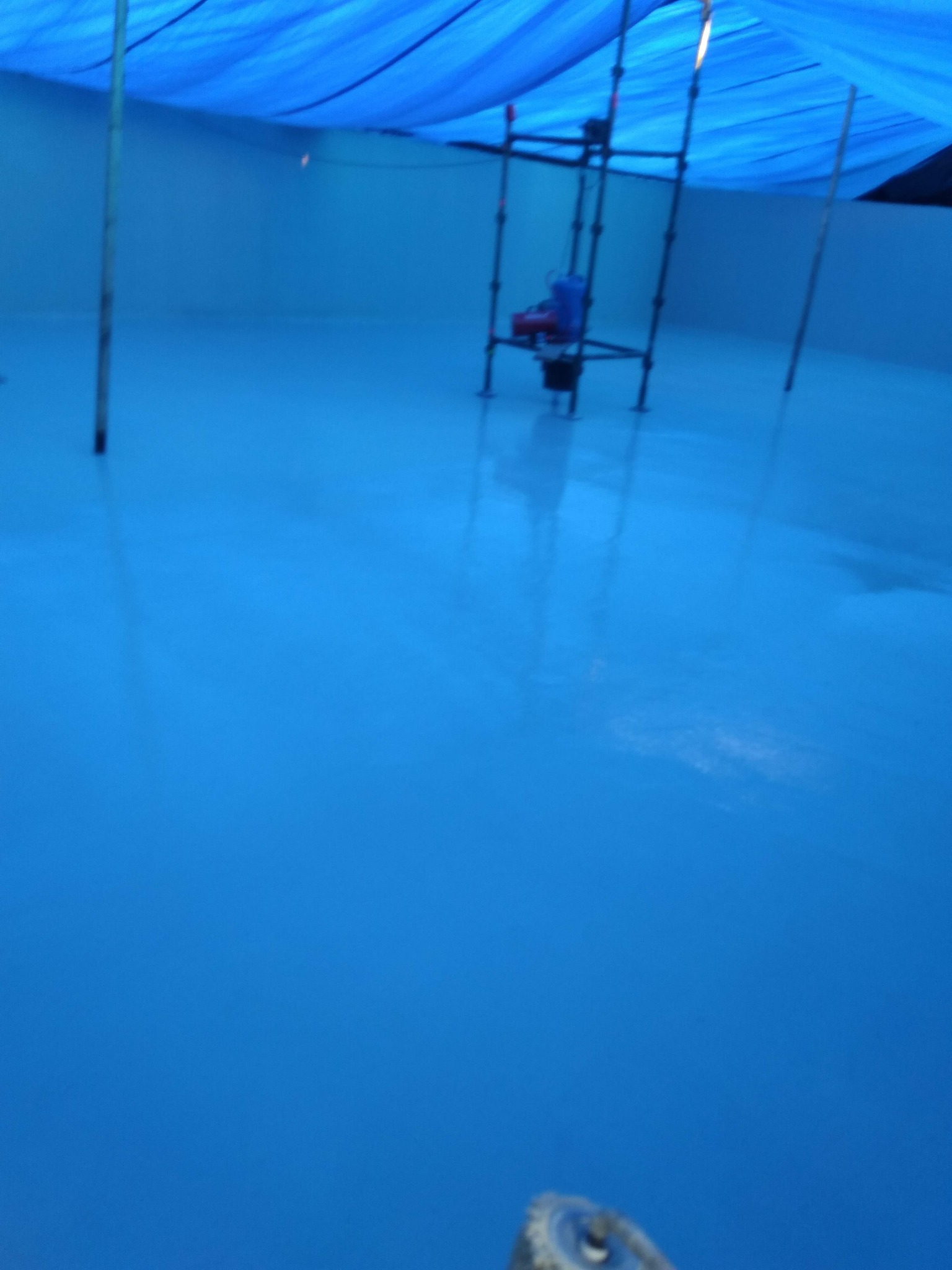 Świeżo wylana, błękitna posadzka w pomieszczeniu z prowizorycznym zadaszeniem z plandeki i metalowym rusztowaniem w tle.