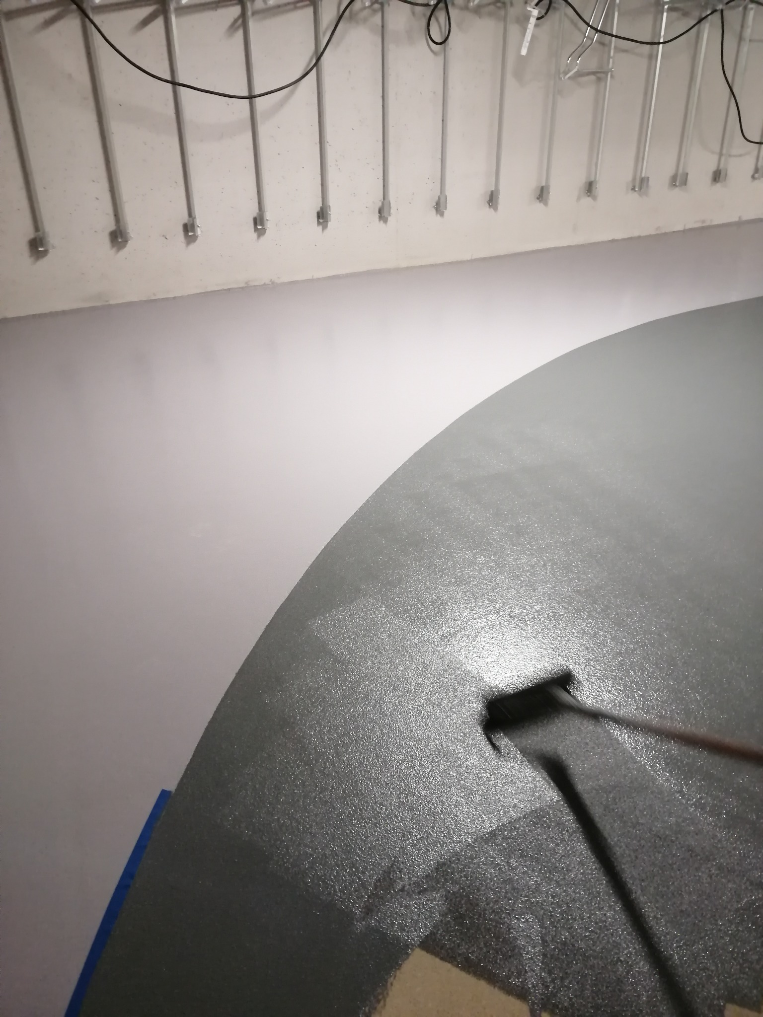 Aplikacja szarej powłoki na posadzce za pomocą wałka, widoczne rury instalacyjne pod sufitem.