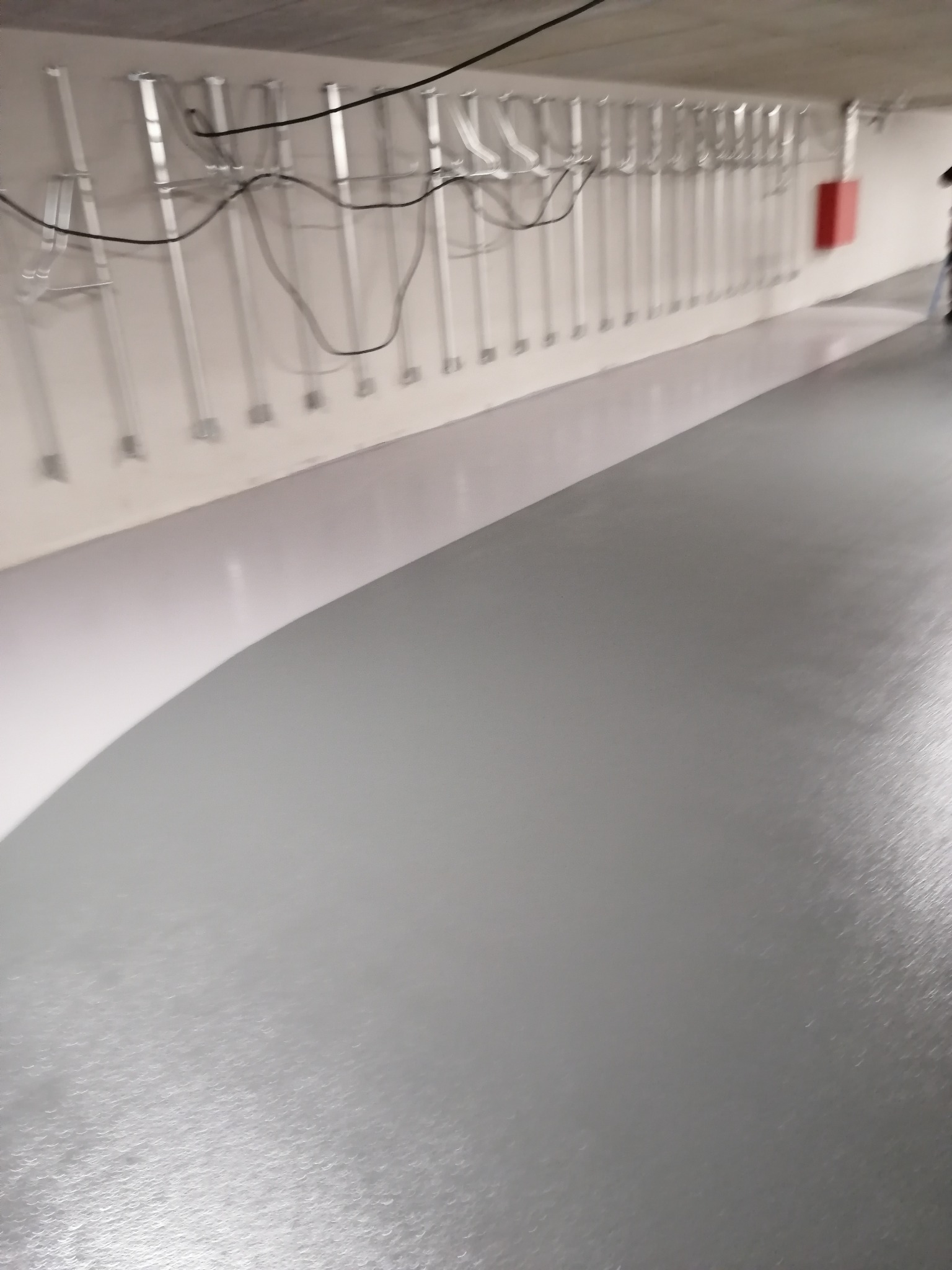 Szara, błyszcząca posadzka w hali magazynowej z widoczną instalacją pod stropem i fragmentem osoby w prawym górnym rogu.