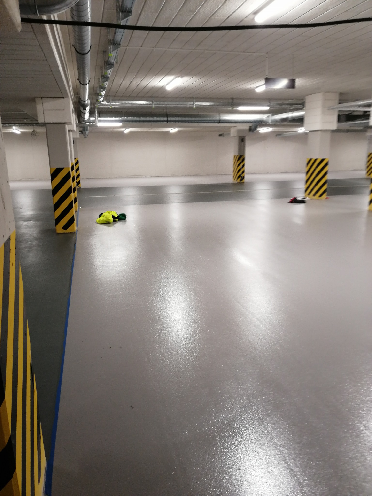 Gładka, szara posadzka w garażu podziemnym, z żółto-czarnymi pasami na filarach i leżącym ubraniem roboczym w tle.