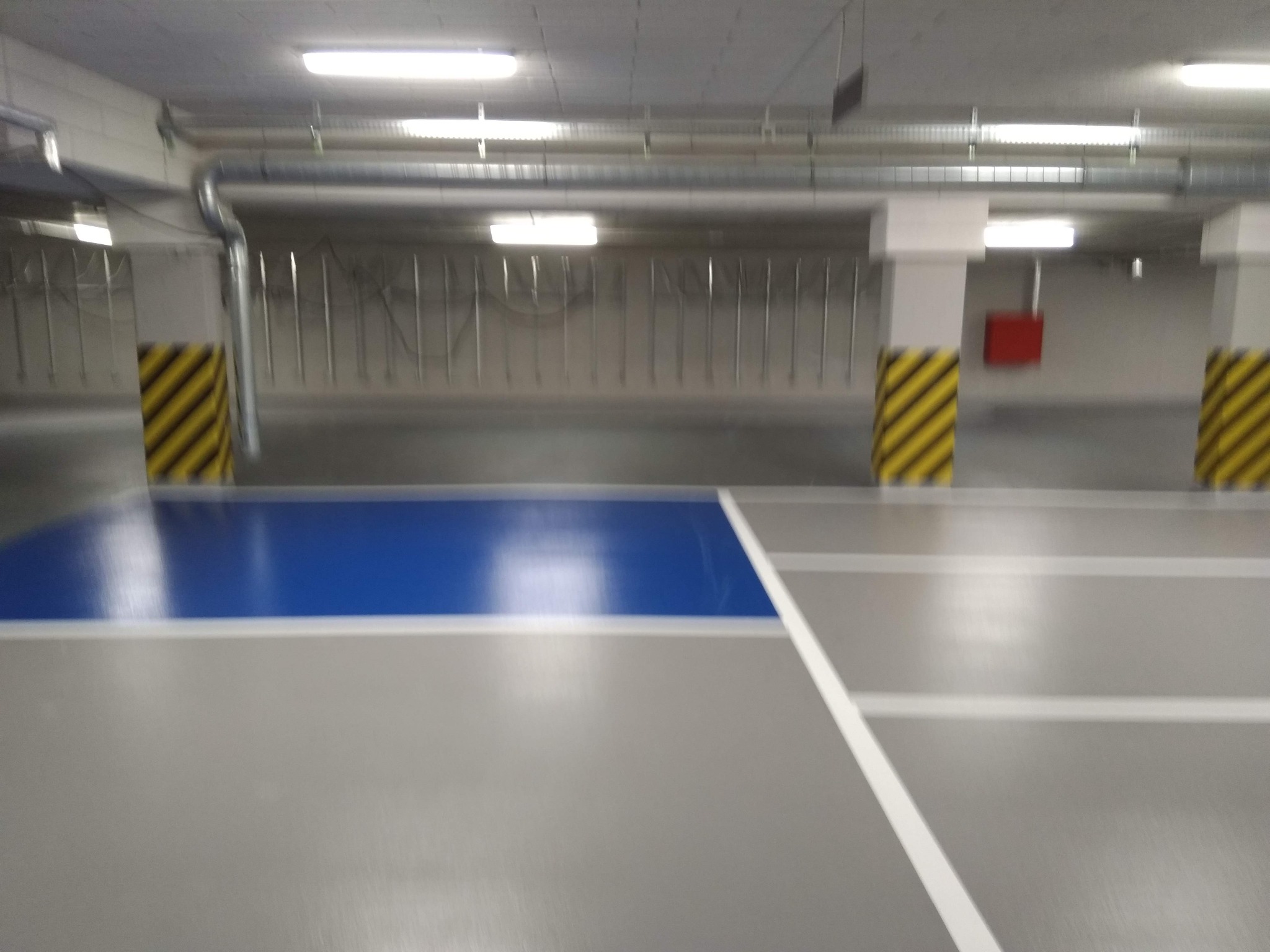 Posadzka żywiczna w garażu podziemnym z wyznaczonym niebieskim miejscem parkingowym i żółto-czarnymi pasami na słupach.