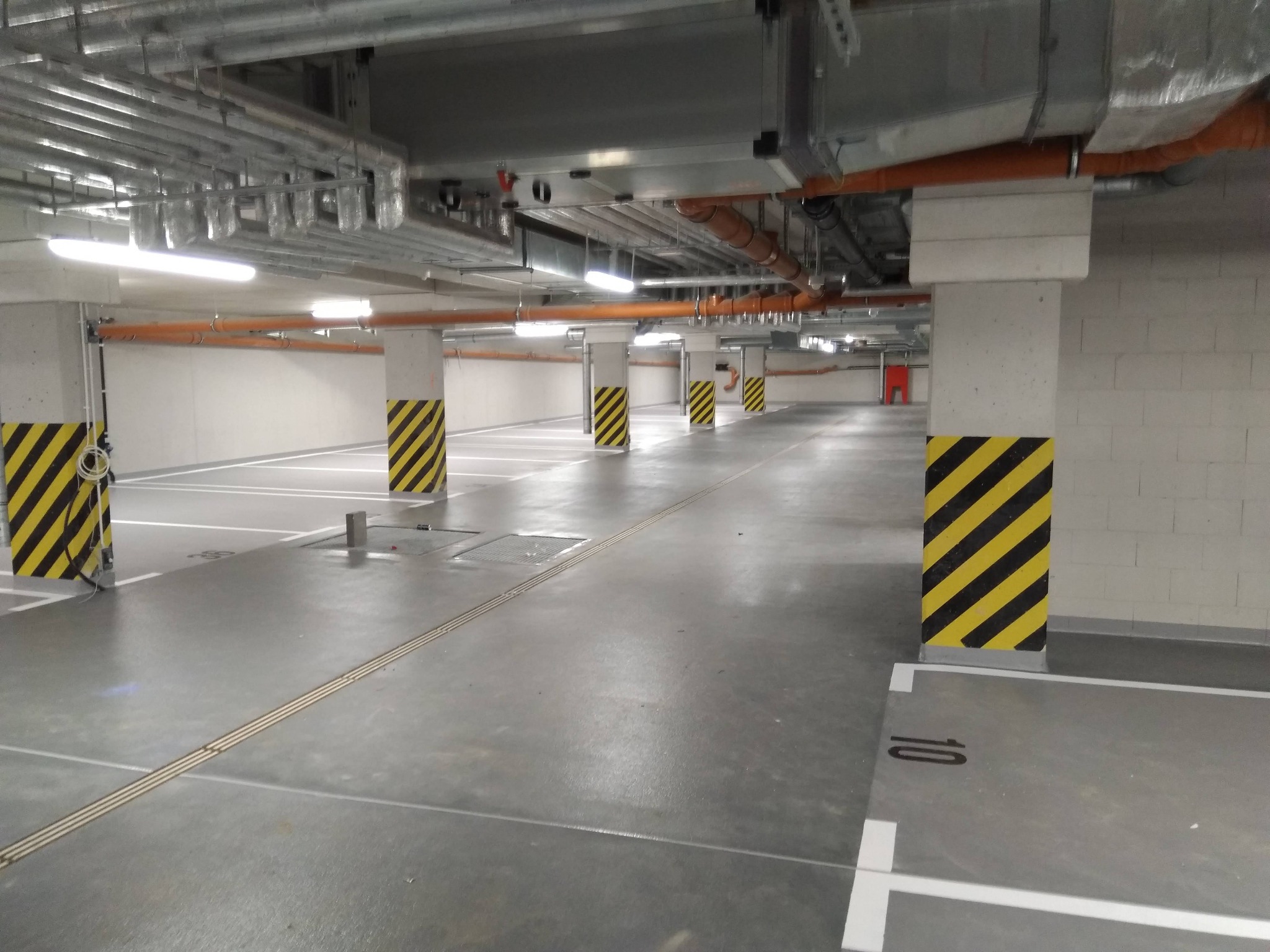 Szary parking podziemny z wyznaczonymi miejscami postojowymi, kolumny oznaczone czarno-żółtymi pasami, instalacje pod sufitem.