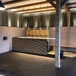 Wnętrze w trakcie prac wykończeniowych: drewniana podłoga, ściany z drewnianymi panelami, nowoczesne oświetlenie wiszące, zabudowa barowa z poziomymi listwami, widoczny słup konstrukcyjny i sprzęt...