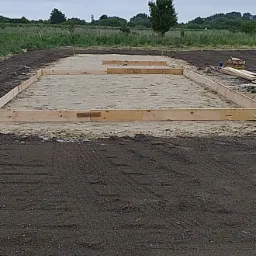 Przygotowany teren pod budowę z drewnianą ramą szalunkową wypełnioną piaskiem, widok z góry na tle pola i drzew w pochmurny dzień.