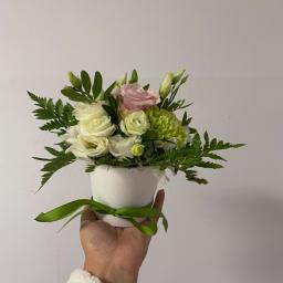Kwiaciarnia Daria Łopacińska - Dłoń w futrzanym mankiecie trzyma biały ceramiczny wazon z kompozycją kwiatową z białych i różowych róż oraz zielonych goździków, przewiązany zieloną wstążką na tle jasnej ściany.