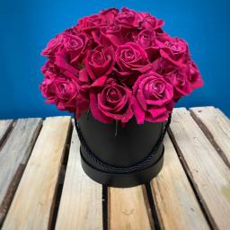 Kwiaciarnia Daria Łopacińska - Bukiet intensywnie różowych róż ułożony w czarnym, okrągłym pudełku z ozdobnym sznurkiem, na tle turkusowej ściany i drewnianego podłoża z desek.