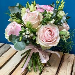 Kwiaciarnia Daria Łopacińska - Delikatny bukiet z różowych róż, goździków i eukaliptusa, przewiązany jasnoróżową wstążką, ułożony na drewnianej palecie na tle niebieskiej ściany.
