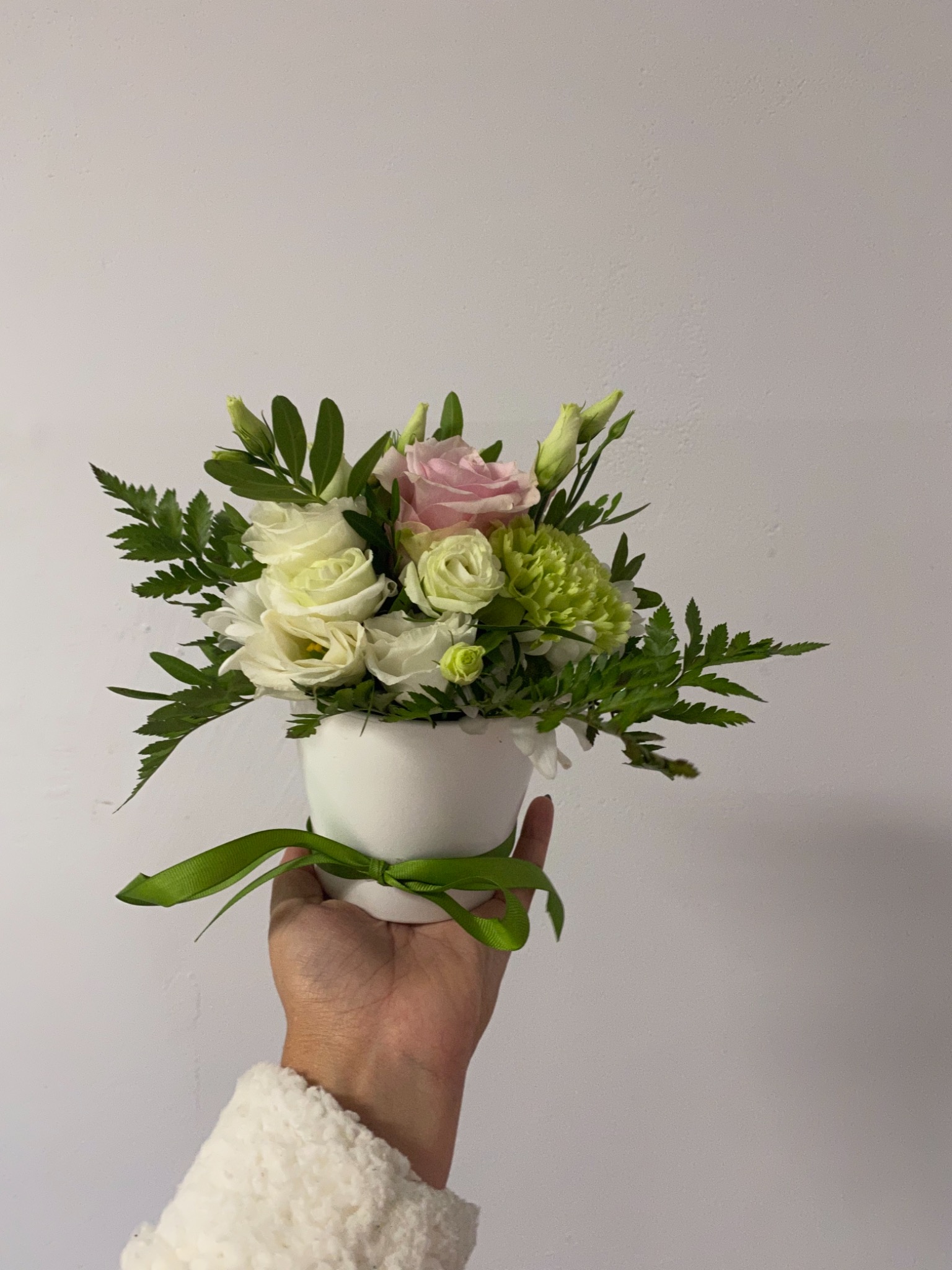 Dłoń w futrzanym mankiecie trzyma biały ceramiczny wazon z kompozycją kwiatową z białych i różowych róż oraz zielonych goździków, przewiązany zieloną wstążką na tle jasnej ściany.
