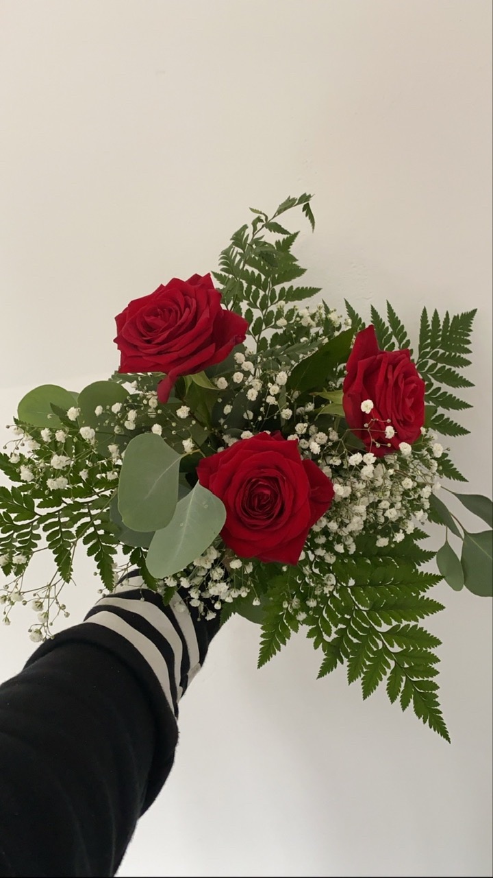 Bukiet z trzech czerwonych róż, paproci i gipsówki trzymany w dłoni ubranej w bluzkę w czarno-białe paski na jasnym tle.