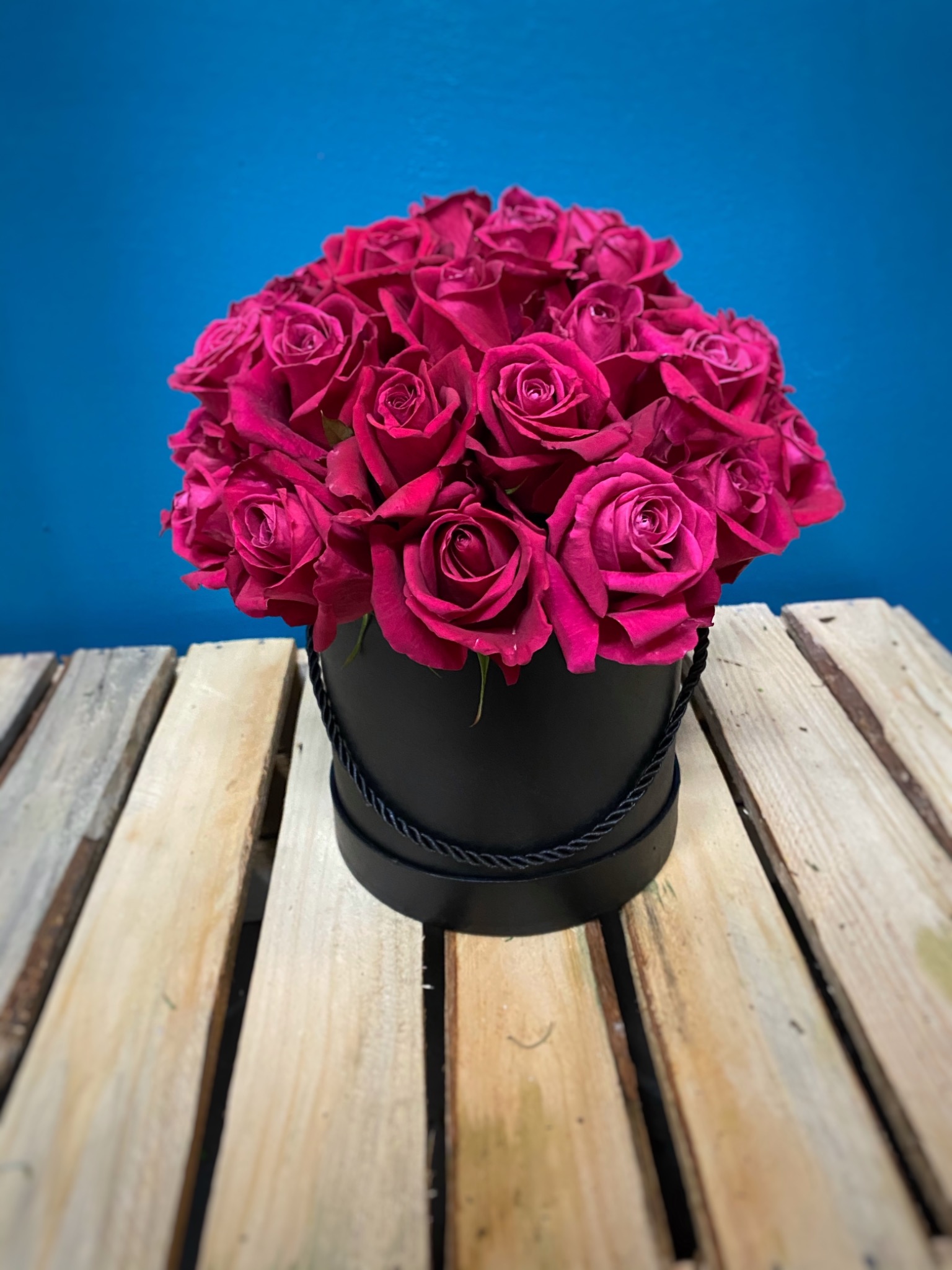 Bukiet intensywnie różowych róż ułożony w czarnym, okrągłym pudełku z ozdobnym sznurkiem, na tle turkusowej ściany i drewnianego podłoża z desek.