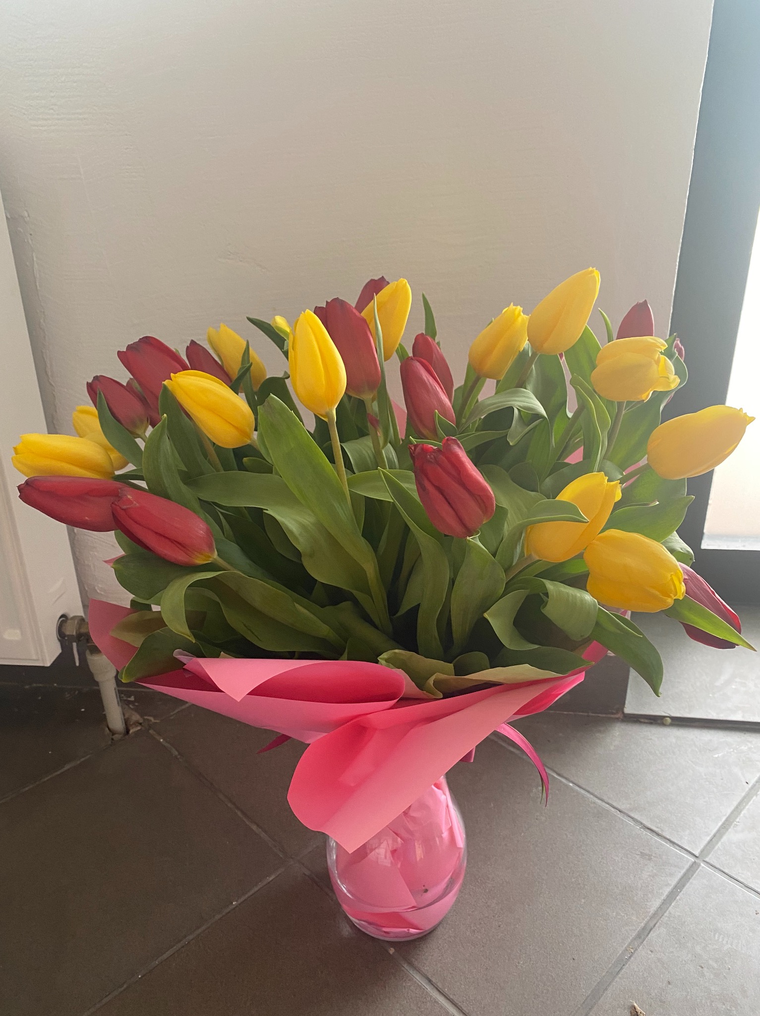 Bukiet czerwonych i żółtych tulipanów w przezroczystym wazonie, owinięty różowym papierem, stojący na ciemnej podłodze w pomieszczeniu.