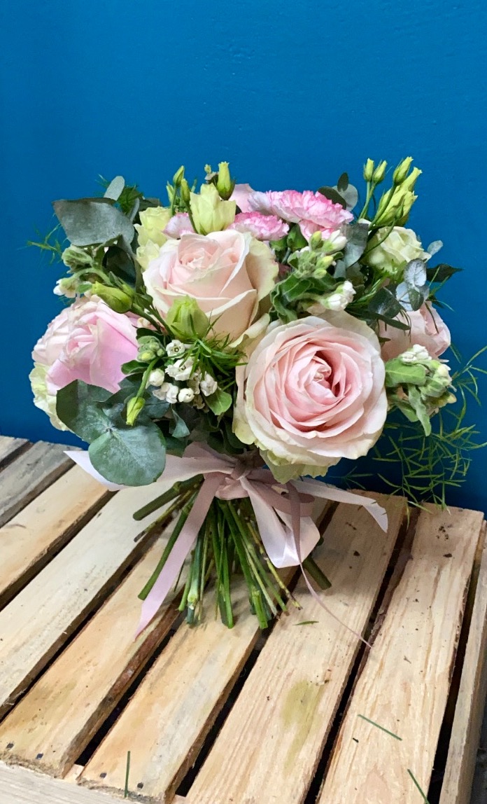 Delikatny bukiet z różowych róż, goździków i eukaliptusa, przewiązany jasnoróżową wstążką, ułożony na drewnianej palecie na tle niebieskiej ściany.
