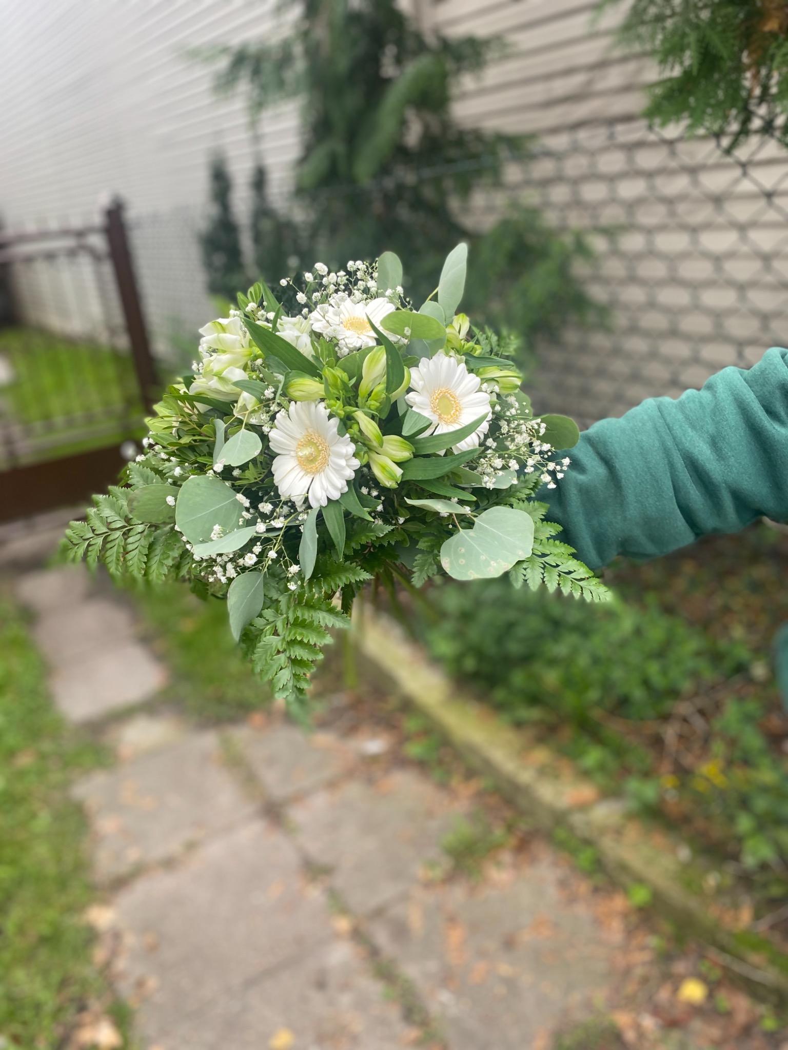 Biało-zielony bukiet z gerberami i eukaliptusem trzymany w dłoni w zielonej bluzie na tle ścieżki i ogrodzenia.