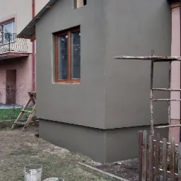 Świeżo otynkowana elewacja budynku z drewnianą ramą okienną, widoczne wiadra z zaprawą i drabina oparta o ścianę, na tle sąsiednich budynków.