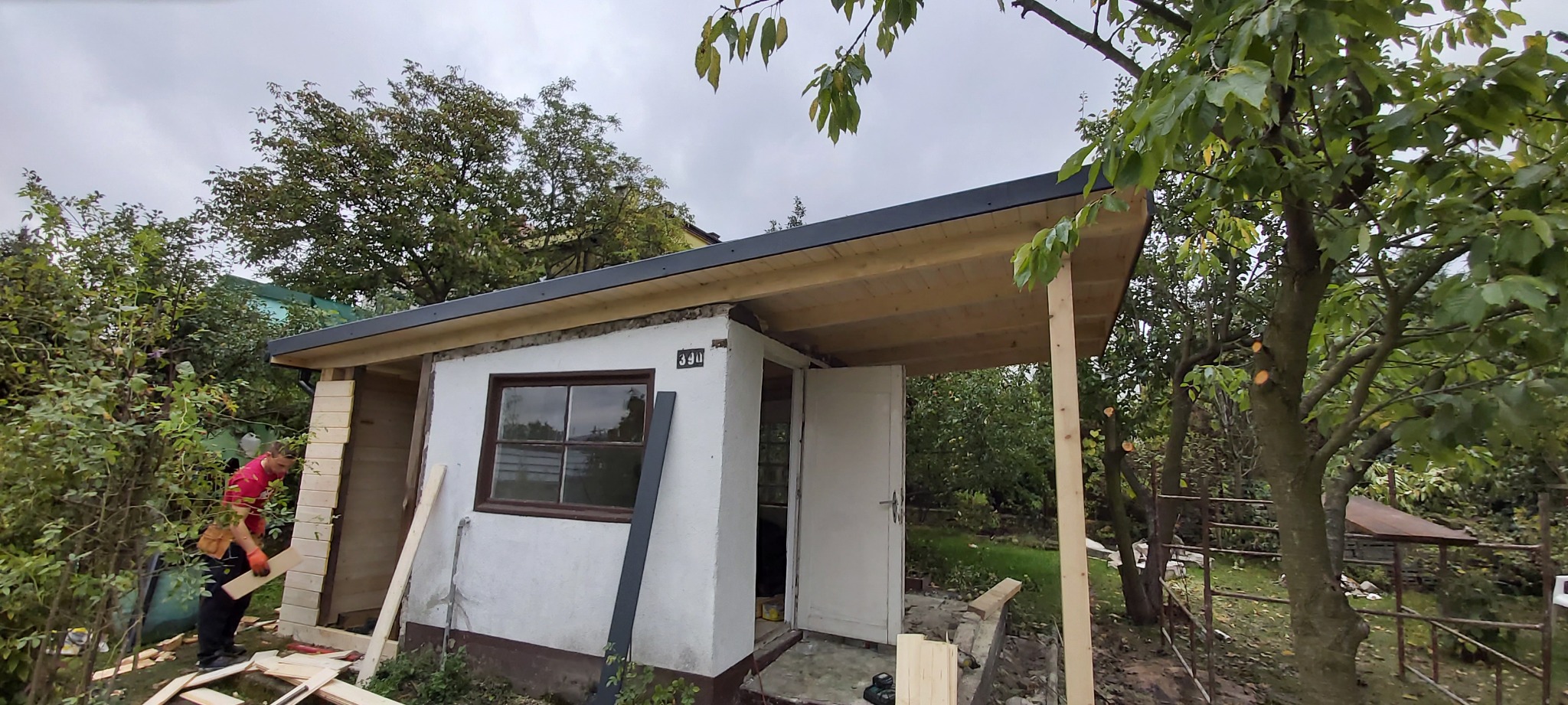 Pracownik w czerwonej koszulce montuje drewniane panele na ścianie zewnętrznej małego budynku z oknem i dwuskrzydłowymi drzwiami, widoczne narzędzia i materiały budowlane, na tle zielony ogród.