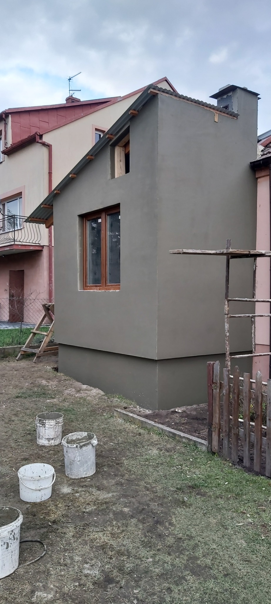 Świeżo otynkowana elewacja budynku z drewnianą ramą okienną, widoczne wiadra z zaprawą i drabina oparta o ścianę, na tle sąsiednich budynków.