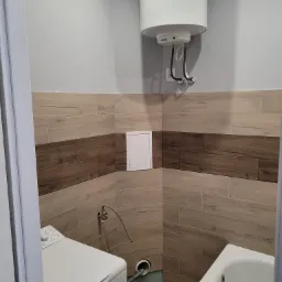 Wykończona łazienka z drewnopodobnymi płytkami na ścianach, białym bojlerem elektrycznym, pralką i wanną, widok zza framugi drzwi.