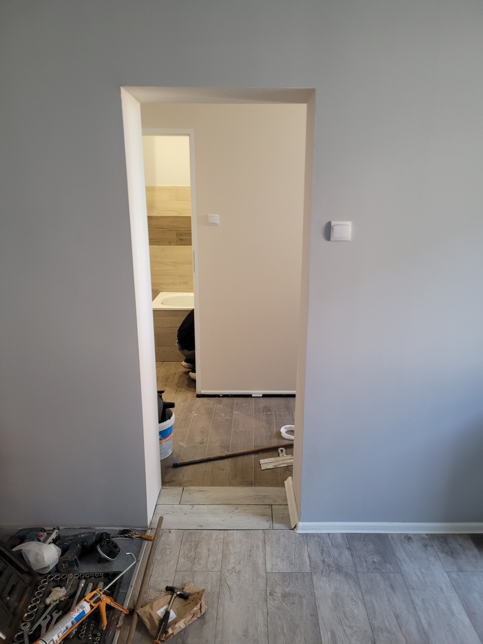 Widok na remontowaną łazienkę z szarymi ścianami, widoczne narzędzia glazurnicze i płytki drewnopodobne na podłodze i ścianach w łazience.