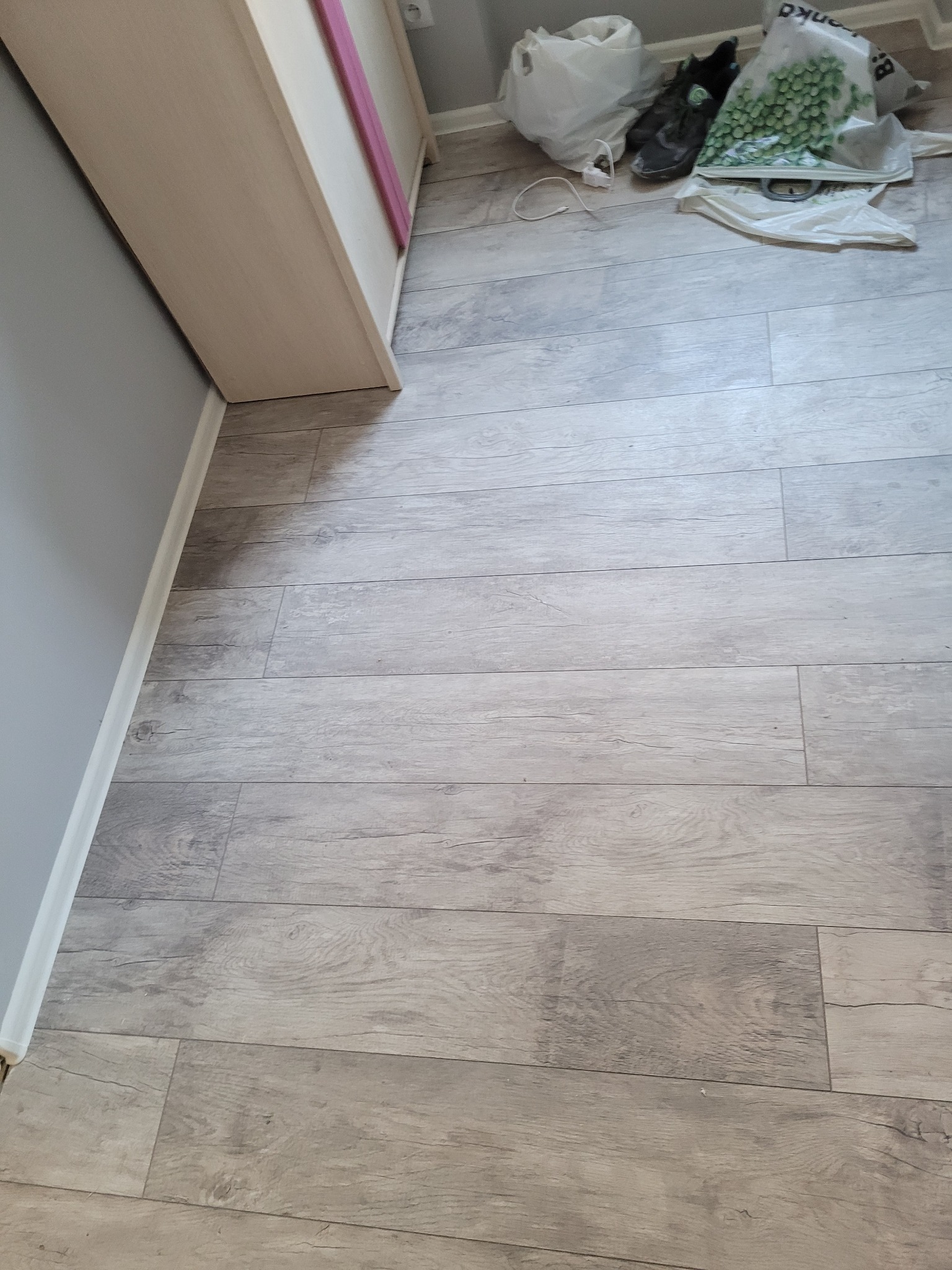 Ułożona podłoga z paneli imitujących drewno w odcieniach szarości, widoczna listwa przypodłogowa, fragment szafy i torby z zakupami w tle.