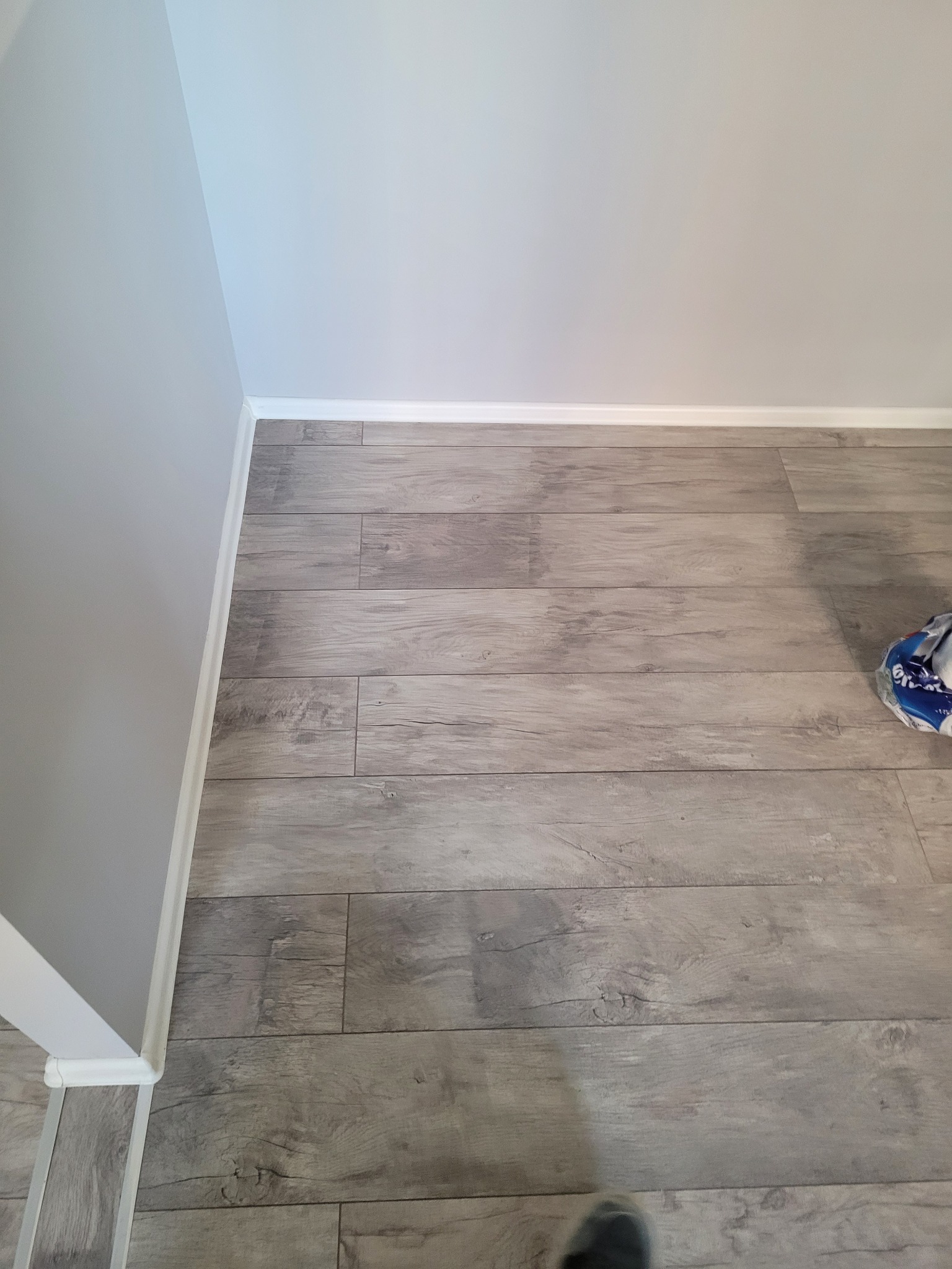 Ułożona podłoga z paneli imitujących drewno w odcieniach szarości, widoczne białe listwy przypodłogowe i fragment ściany pomalowanej na szaro, w rogu leży opakowanie po produkcie.