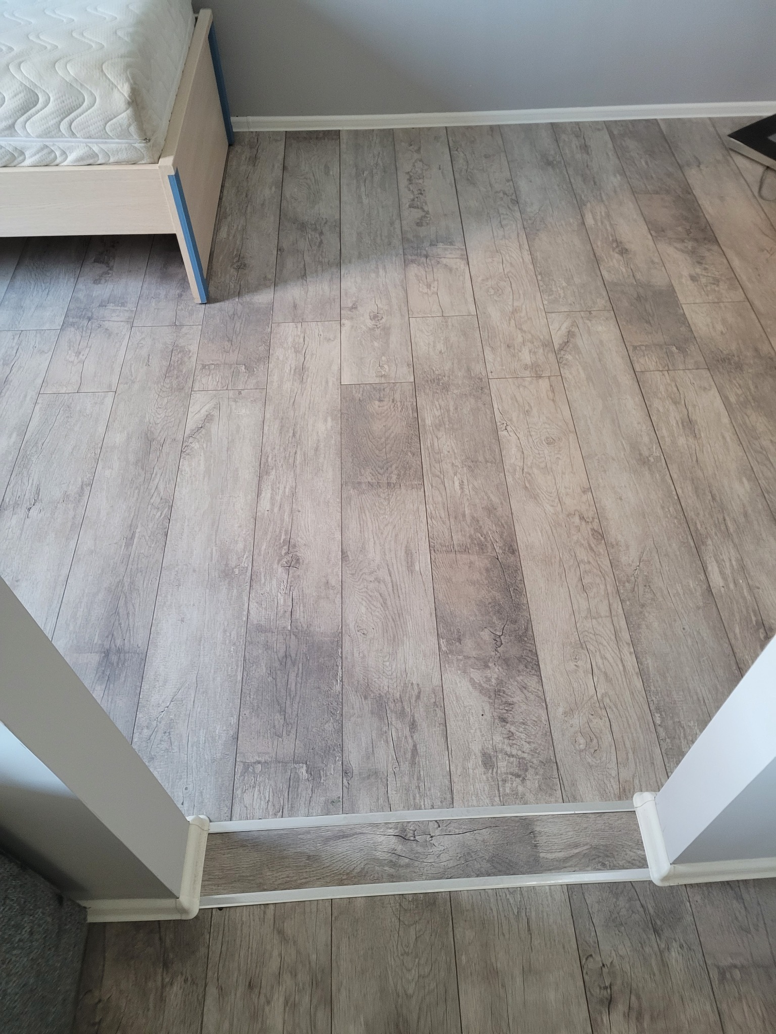 Ułożona podłoga z paneli imitujących jasne drewno w pokoju, widoczny fragment łóżka i próg drzwiowy z listwą.