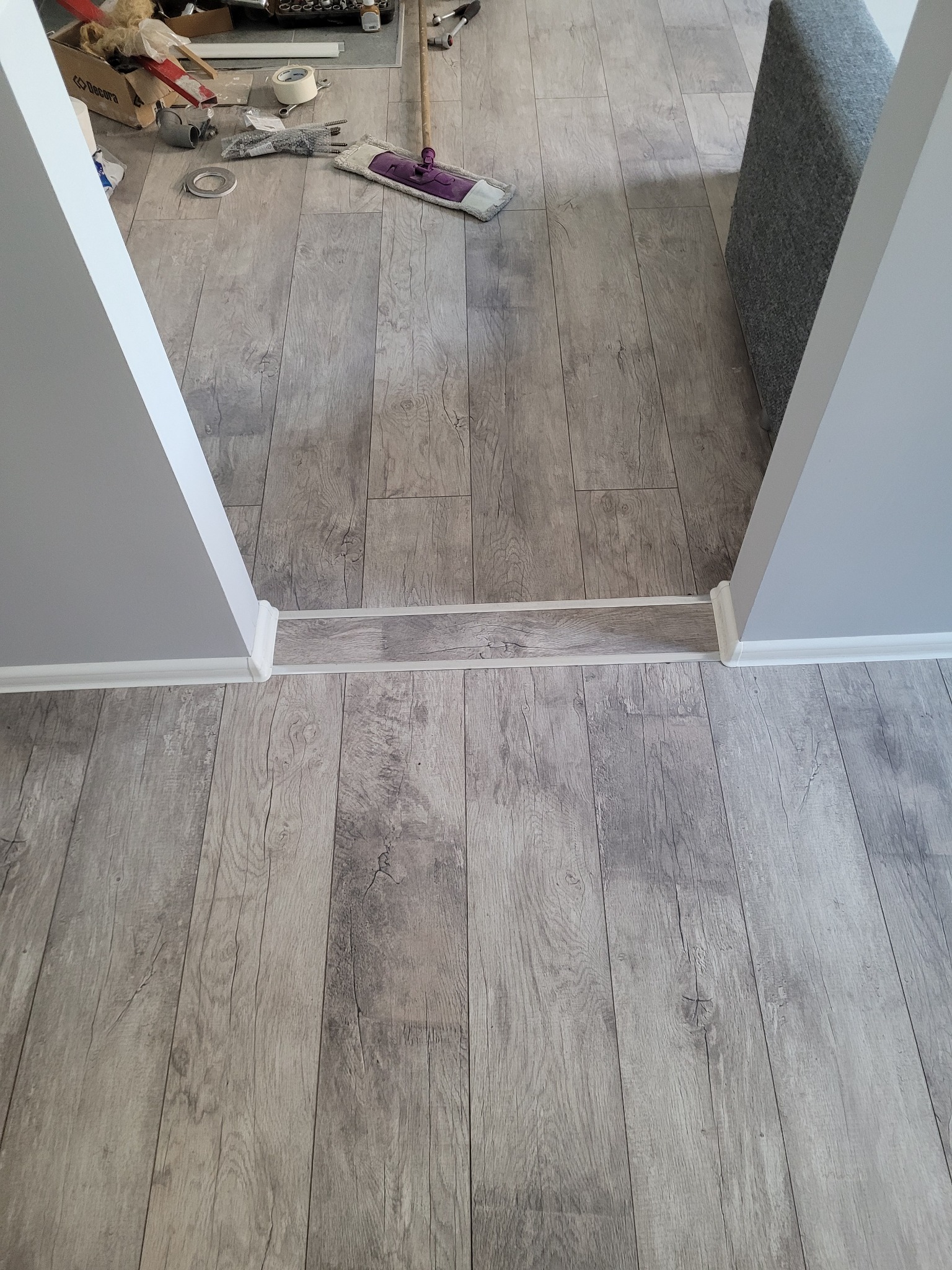 Ułożona podłoga z paneli imitujących drewno w odcieniach szarości, widoczne narzędzia i akcesoria glazurnicze w tle, listwy przypodłogowe przy ścianie.