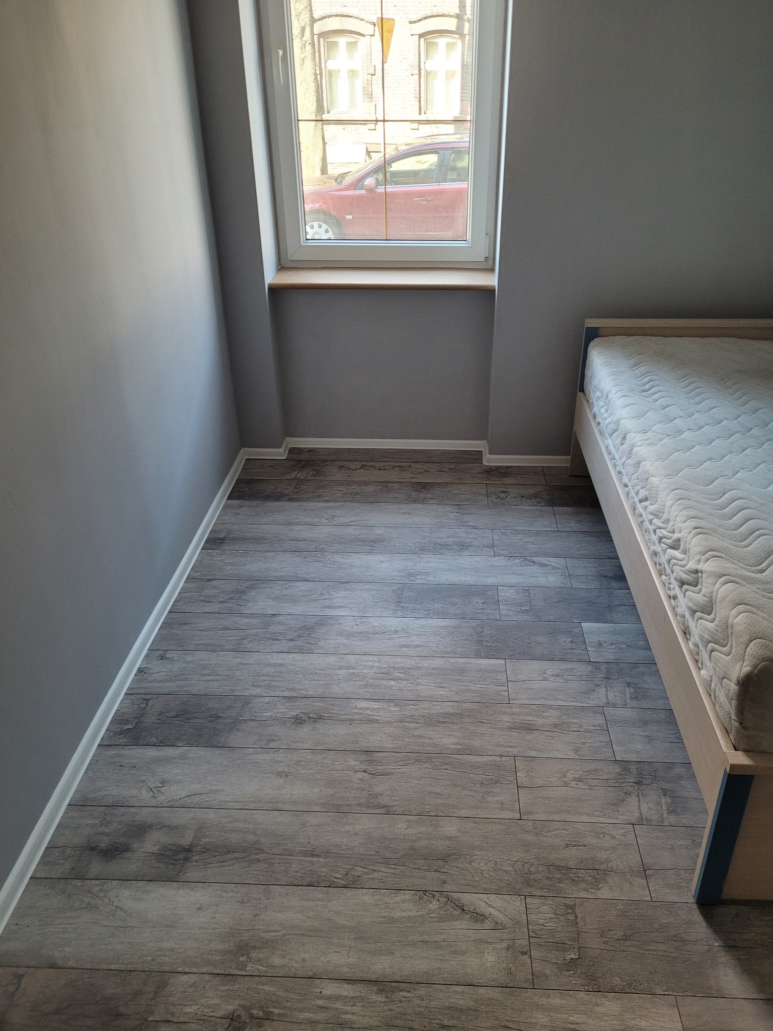 Szare panele podłogowe imitujące drewno ułożone w pokoju w Katowicach, widok na fragment łóżka i okno z widokiem na ulicę.