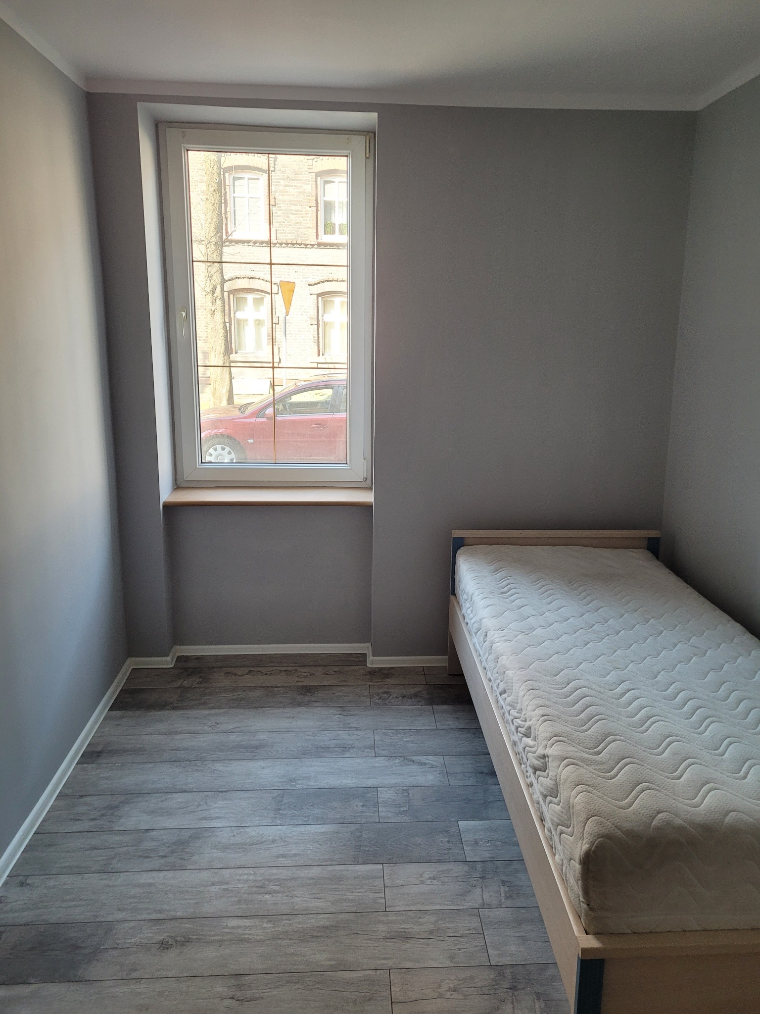 Pokój po remoncie: Szare ściany, szara podłoga imitująca drewno, okno z widokiem na ceglany budynek i czerwony samochód, łóżko jednoosobowe z materacem.