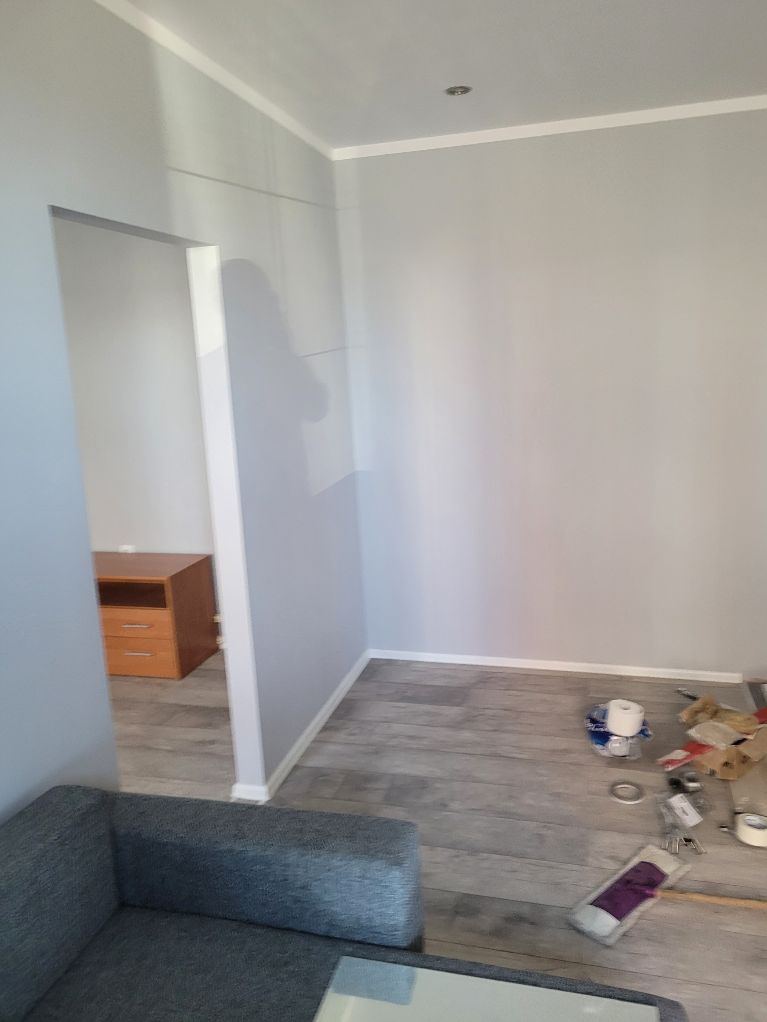 Pokój po malowaniu ścian na szaro, widoczne ślady po pracach malarskich na podłodze i narożnik szarej sofy.