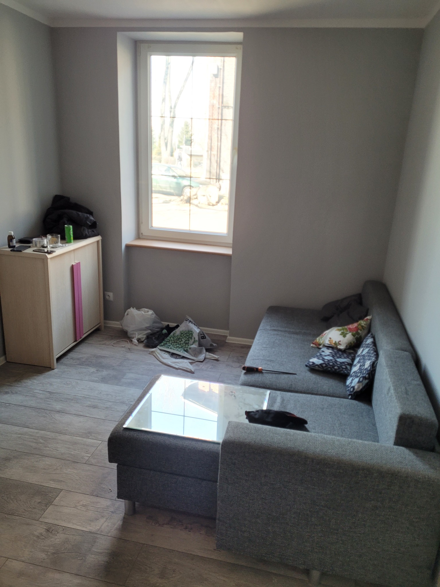 Szare wnętrze pokoju z narożnikiem, stolikiem kawowym ze szklanym blatem i szafką z różowymi akcentami, w trakcie drobnych prac wykończeniowych lub porządkowych.