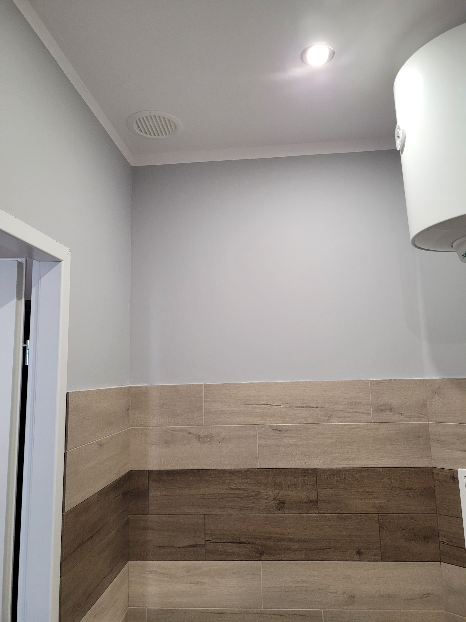 Świeżo wykończona łazienka z drewnopodobnymi płytkami ułożonymi w pasy, widoczny fragment bojlera i kratka wentylacyjna w suficie.