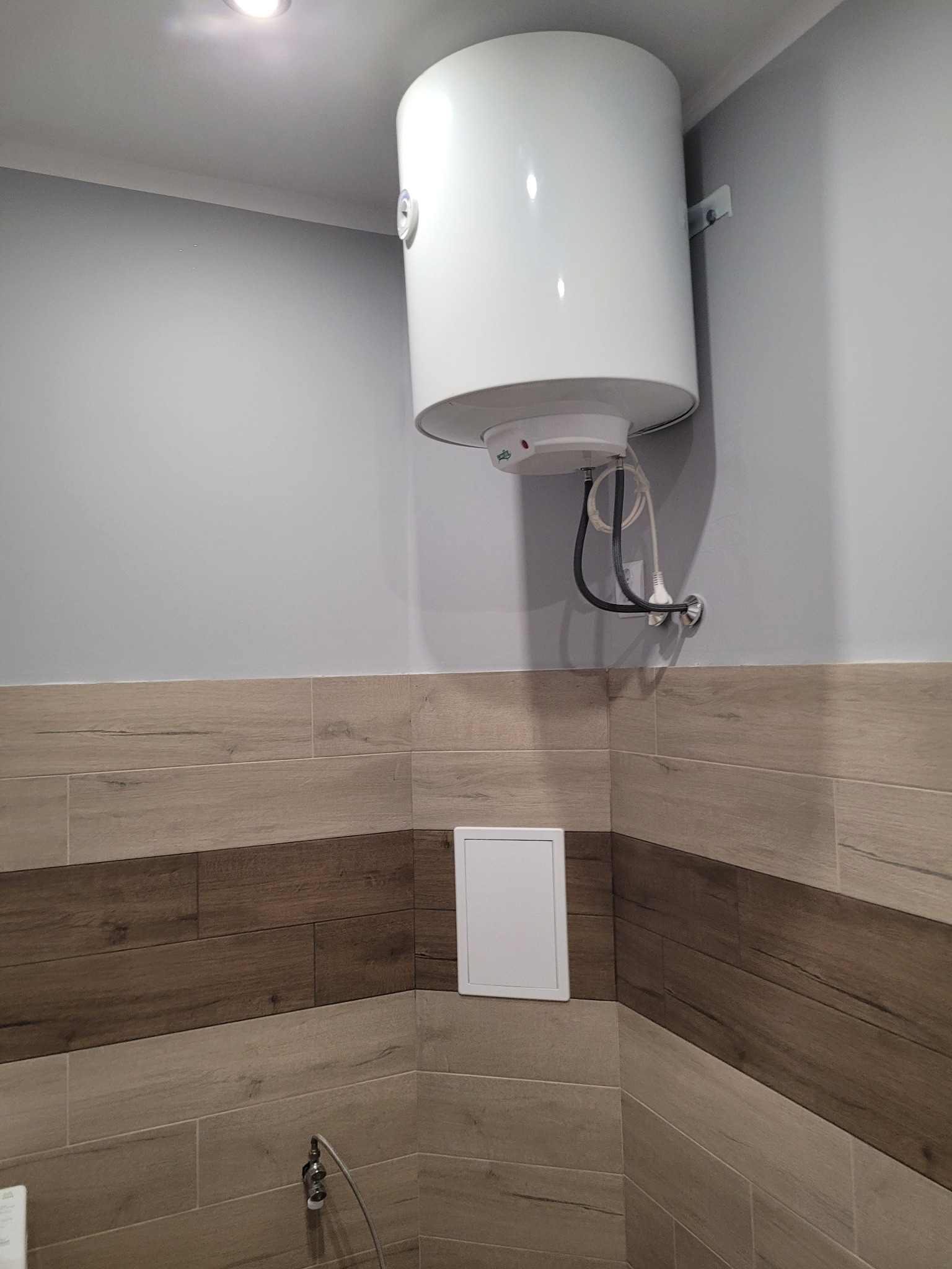 Zamontowany na ścianie, biały bojler elektryczny w łazience wyłożonej płytkami imitującymi drewno, z widocznymi przyłączami hydraulicznymi i elektrycznymi.