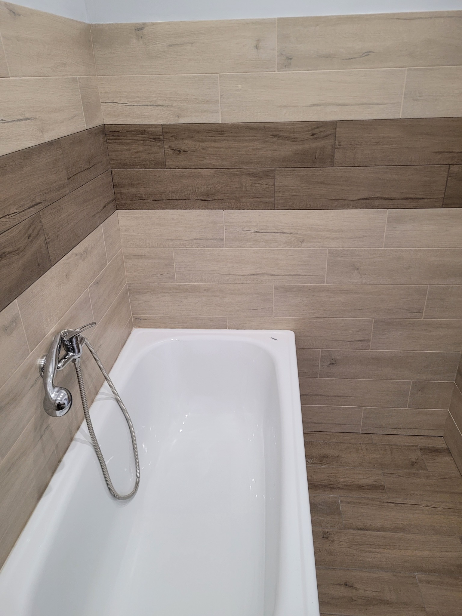 Nowoczesna łazienka z wanną, wykończona płytkami imitującymi drewno w odcieniach beżu i brązu, z chromowaną baterią prysznicową.