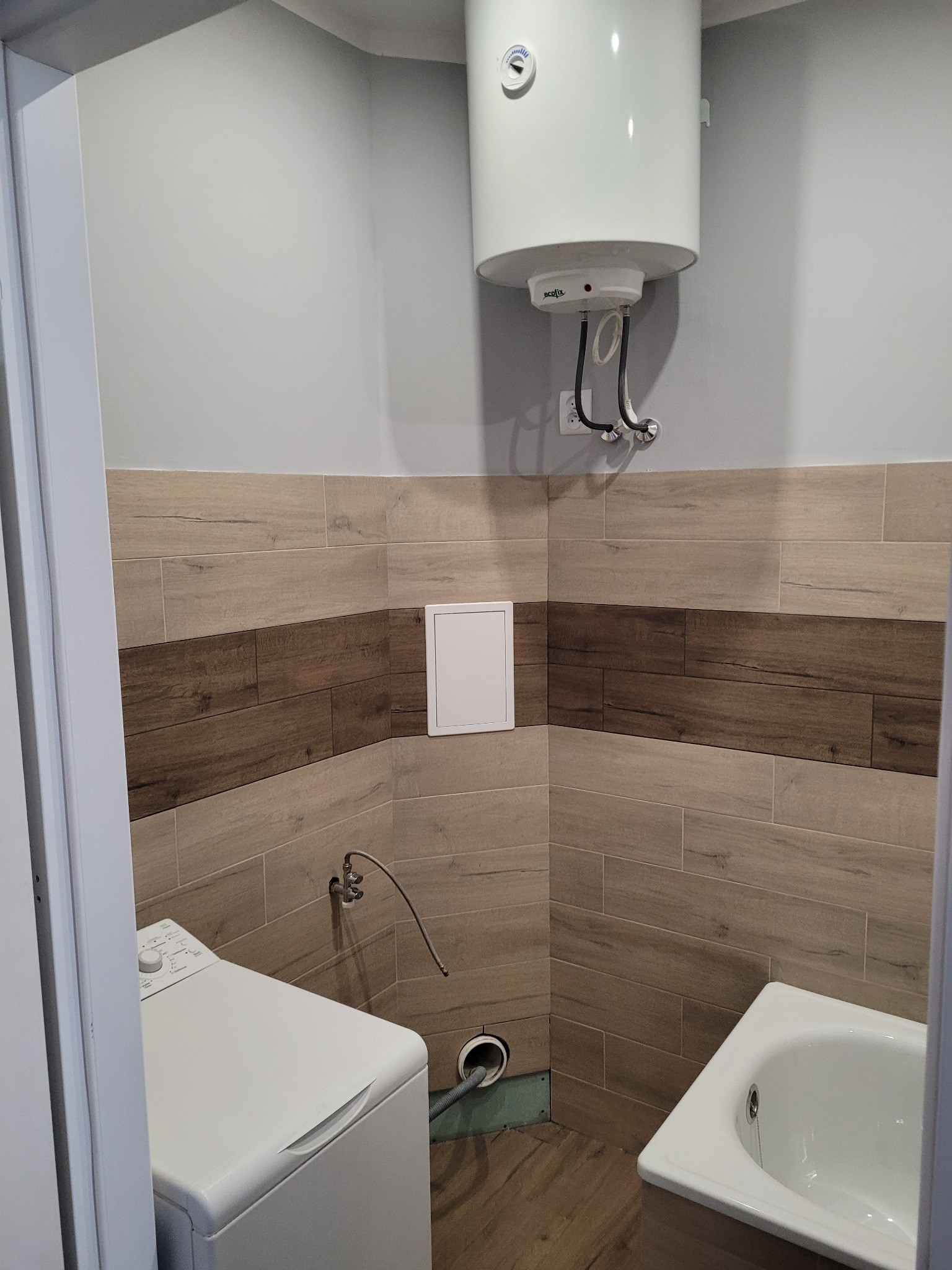 Wykończona łazienka z drewnopodobnymi płytkami na ścianach, białym bojlerem elektrycznym, pralką i wanną, widok zza framugi drzwi.