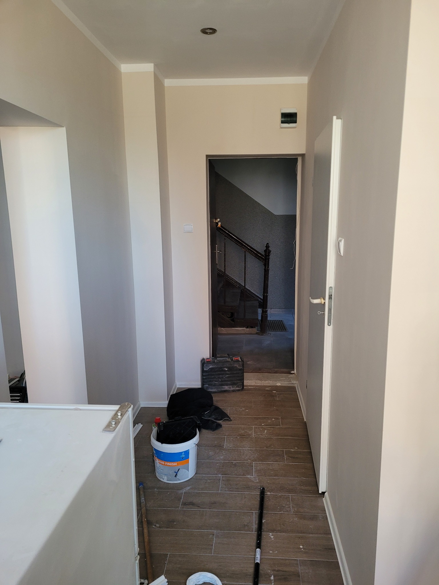 Wnętrze korytarza w trakcie malowania: ściany w odcieniach beżu i szarości, widoczne narzędzia malarskie, wiadro z farbą, podłoga wyłożona płytkami imitującymi drewno, otwarte drzwi prowadzące...